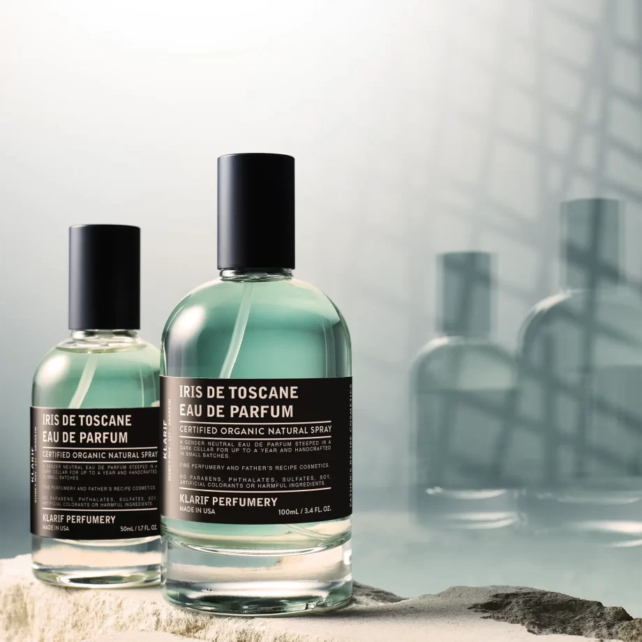 A sleek, clear glass bottle of Klarif Iris de Toscane Eau de Parfum, the Klarif signature scent, with a minimalist design and a black label, set against a backdrop of soft, white rock and subtle green elements, emphasizing its organic and fresh, oceanic inspiration.